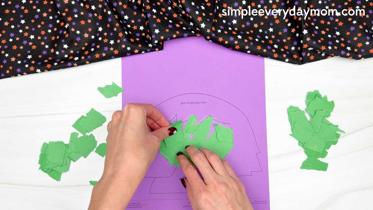 Hands gluing torn green paper pieces onto the witch's face outline on a purple Witch Torn Paper Craft template.