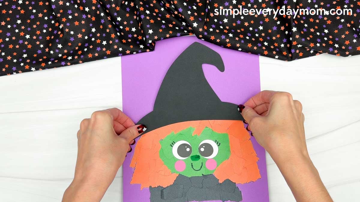 Hands positioning a black witch hat above the green face and orange torn paper hair for the Witch Torn Paper Craft.