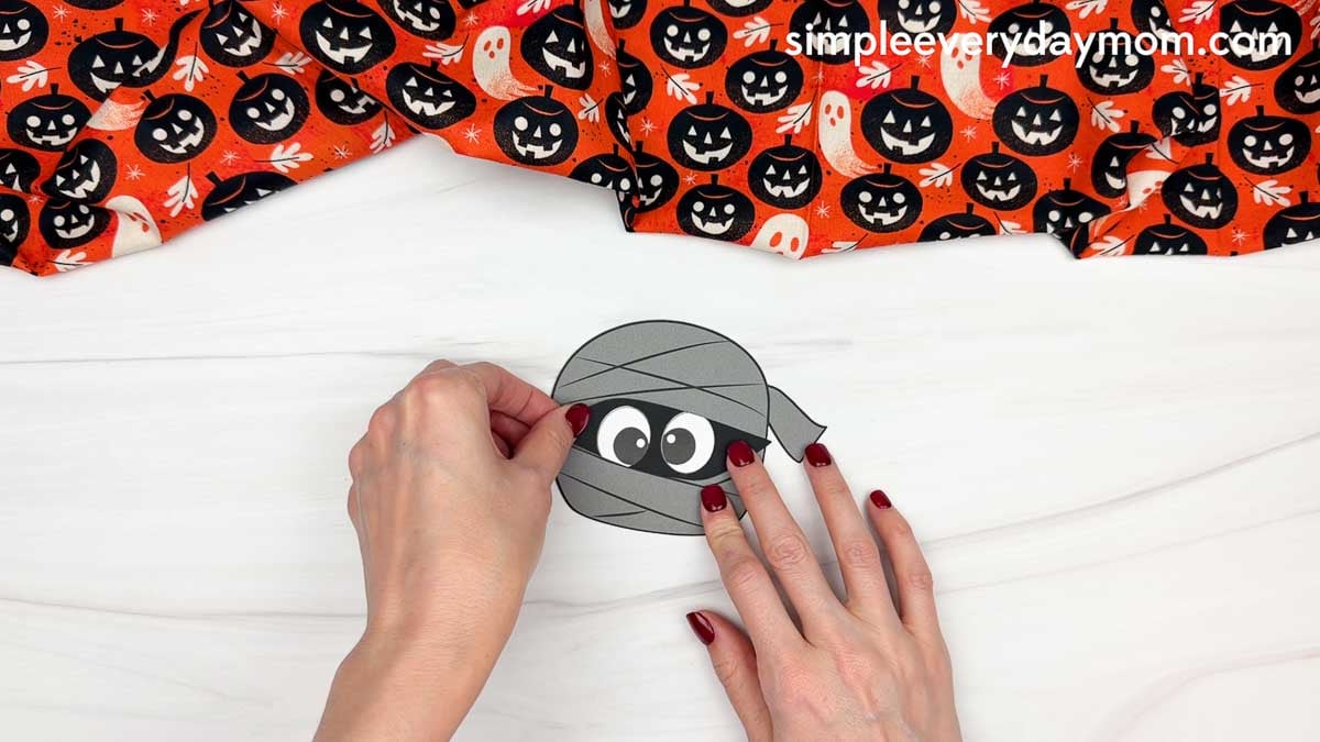 Hands gluing the eyes piece onto the center of the mummy head for a Halloween name craft for kids.