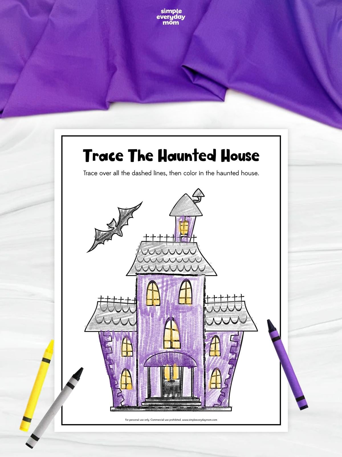 Haunted house tracing and coloring worksheet featuring a completed purple house with gray roofs, yellow windows, and two bats flying above. Crayons in yellow, gray, and purple surround the worksheet.