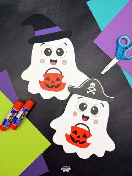 A completed craft display of two Halloween paper ghosts wearing witch and pirate hats and holding pumpkin buckets, surrounded by craft supplies.