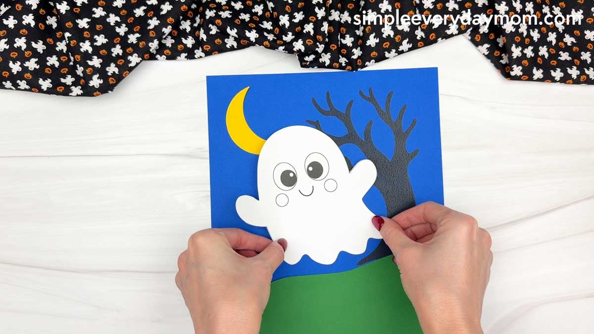Hands positioning the smiling ghost piece over the blue background, crescent moon, and spooky tree for the Halloween Ghost Name Craft.