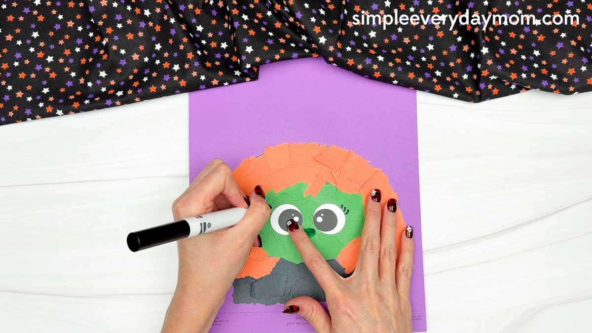 Hands using a black marker to draw eyelashes on the witch's eyes for the Witch Torn Paper Craft.