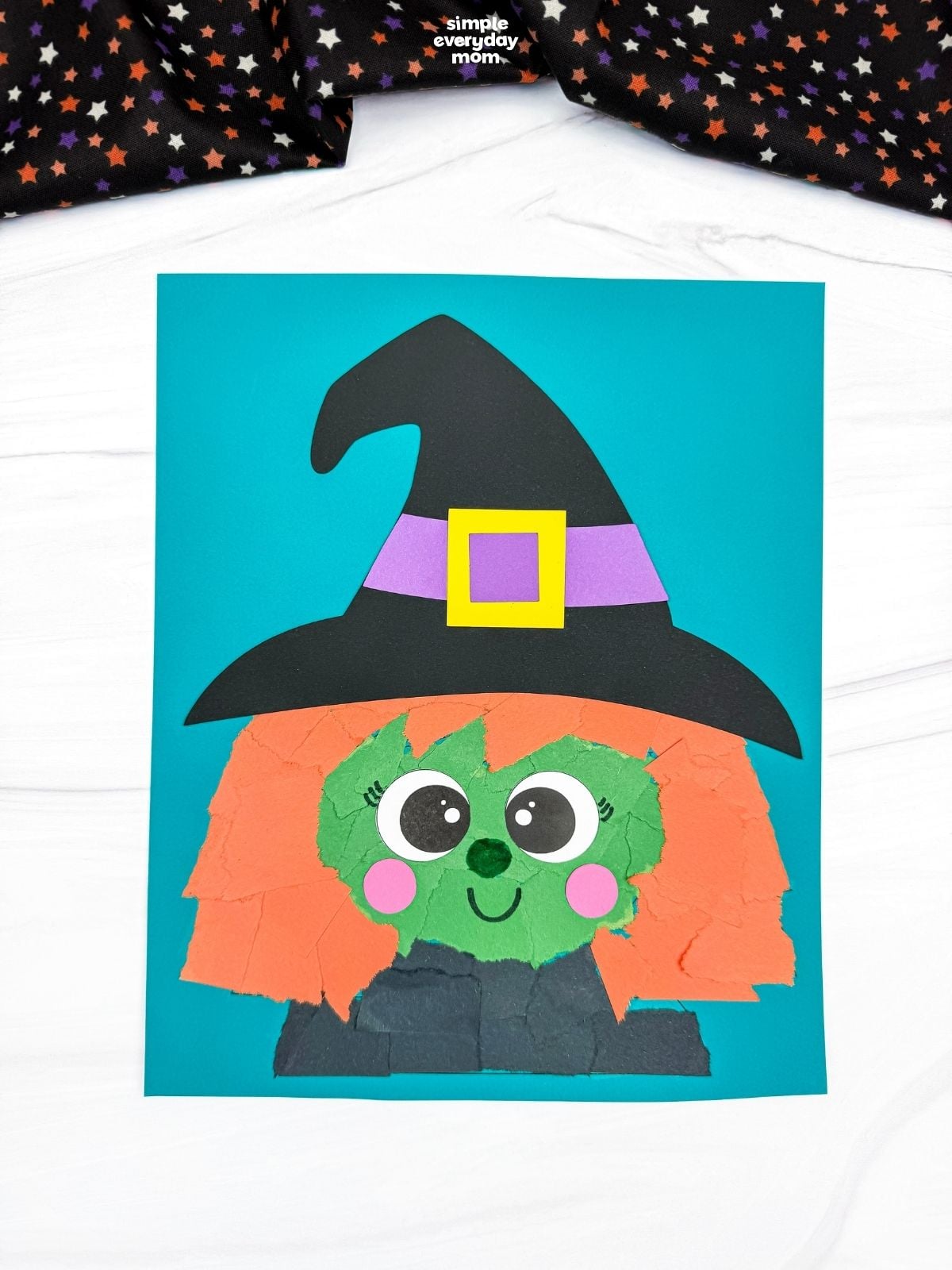 A completed Witch Torn Paper Craft featuring orange torn paper hair, a green face, pink cheeks, and a black witch hat on a blue background.