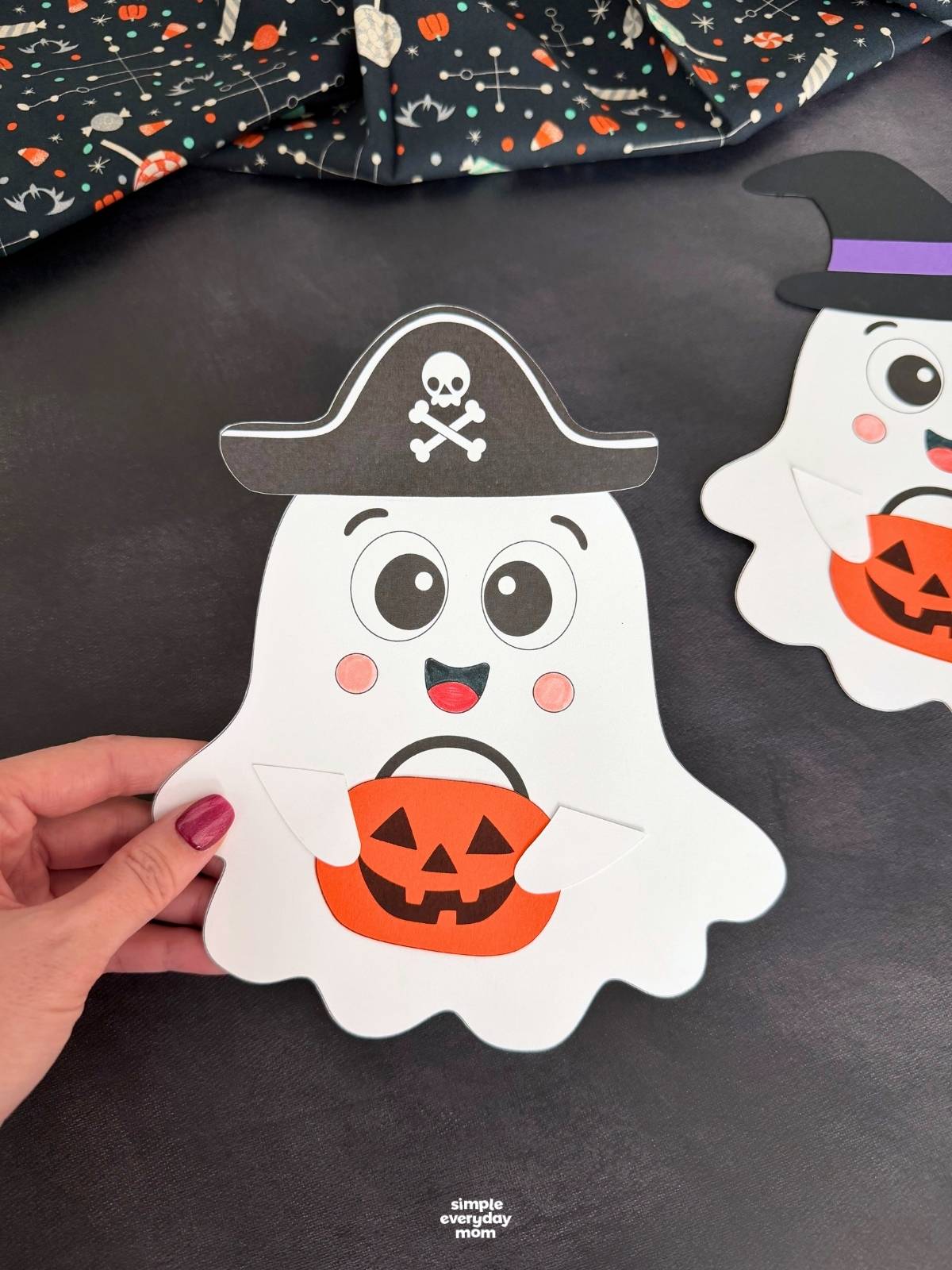 Hands holding a Halloween paper ghost craft wearing a skull-and-crossbones pirate hat and holding an orange pumpkin bucket.