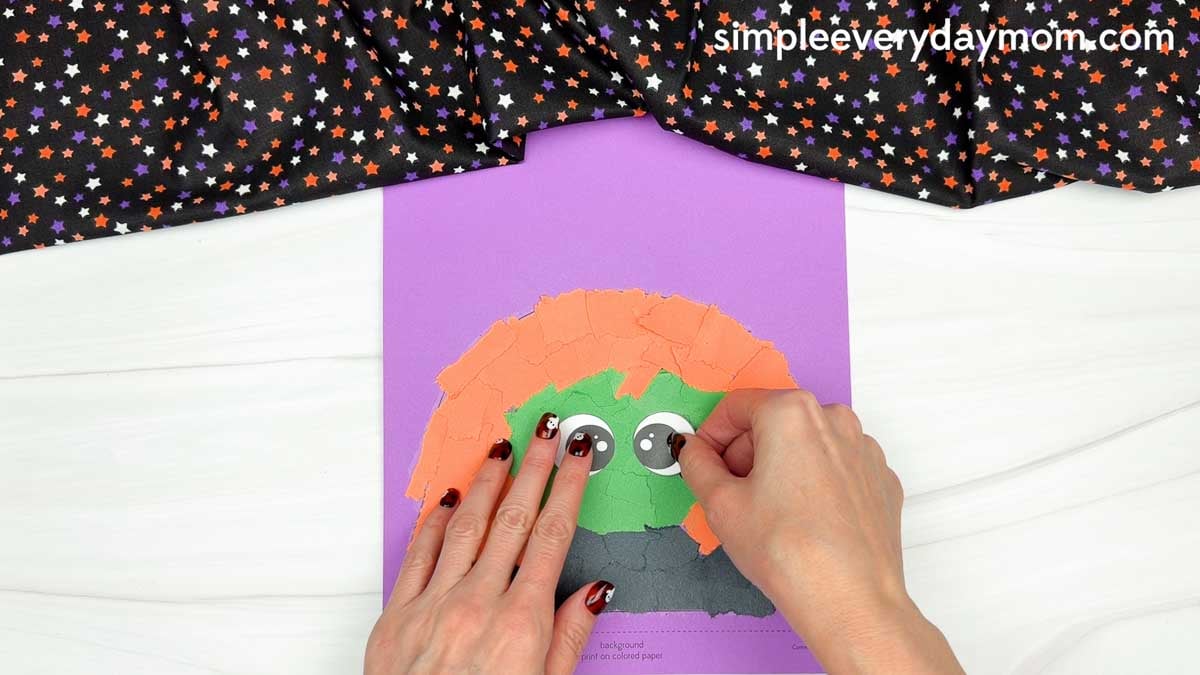 Hands gluing the witch's eyes onto the green face surrounded by torn orange paper hair for the Witch Torn Paper Craft.