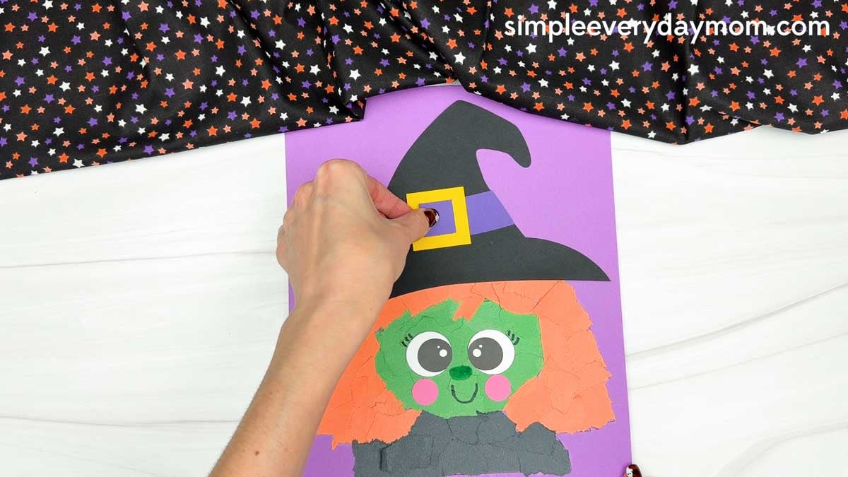 Hands gluing the inner buckle onto the yellow outer buckle of the witch's hat for the Witch Torn Paper Craft.