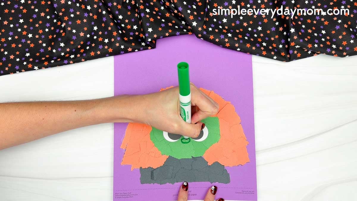 Hands using a green marker to draw the witch's nose on the face of the Witch Torn Paper Craft.