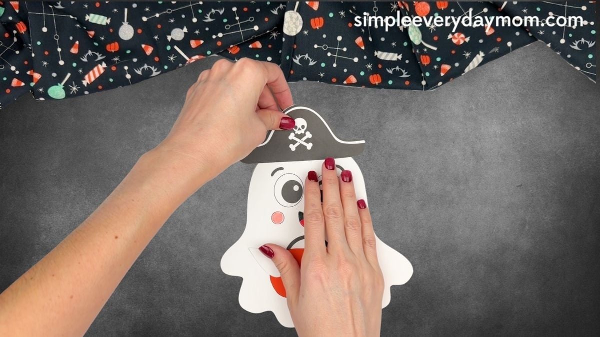 Hands attaching a skull-and-crossbones pirate hat to the top of a Halloween paper ghost craft.
