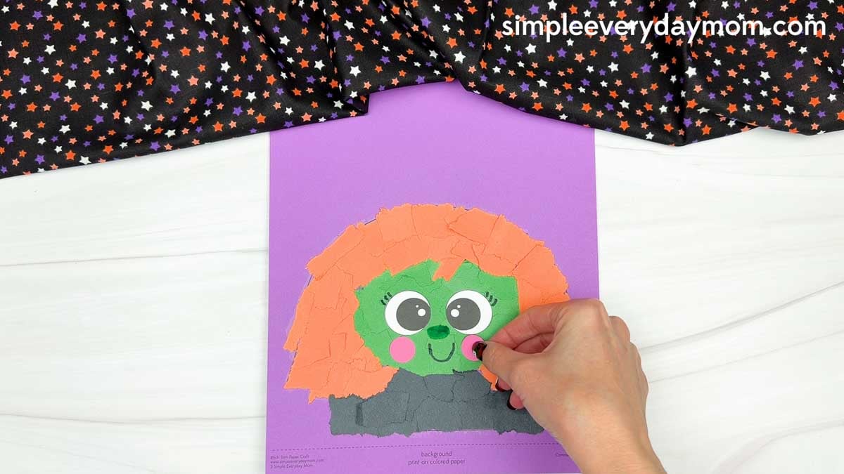 Hands gluing a pink cheek onto the witch's green face with orange torn paper hair for the Witch Torn Paper Craft.