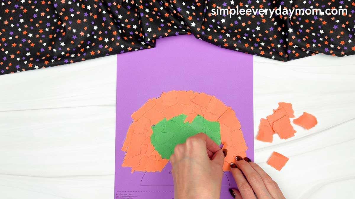 Hands gluing torn orange paper pieces around the green witch's face to create hair for the Witch Torn Paper Craft.