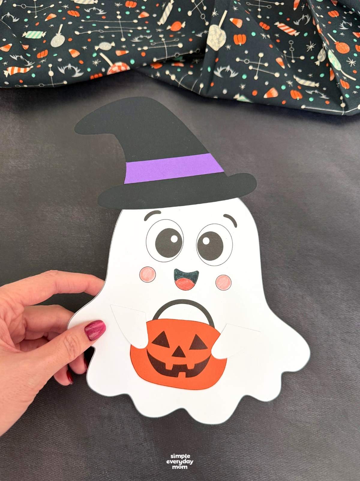 Hands holding a smiling Halloween paper ghost craft wearing a black witch hat with a purple band and holding an orange pumpkin bucket.