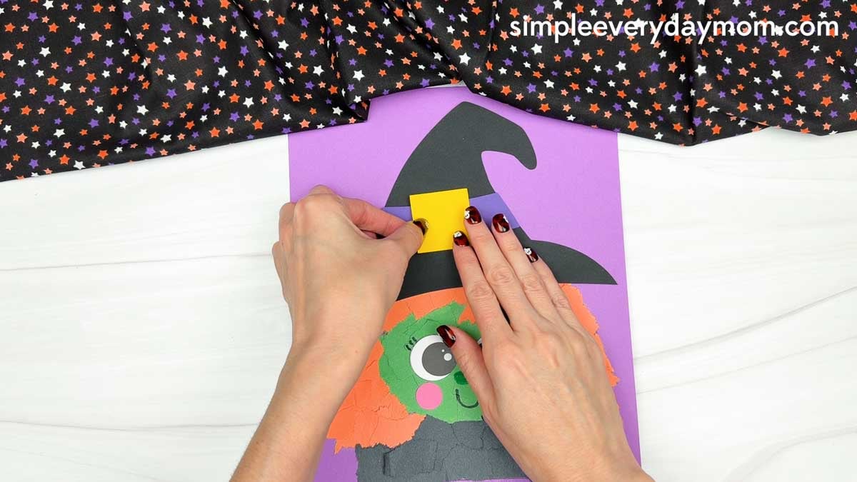 Hands gluing a yellow outer buckle onto the purple hat band of the Witch Torn Paper Craft.