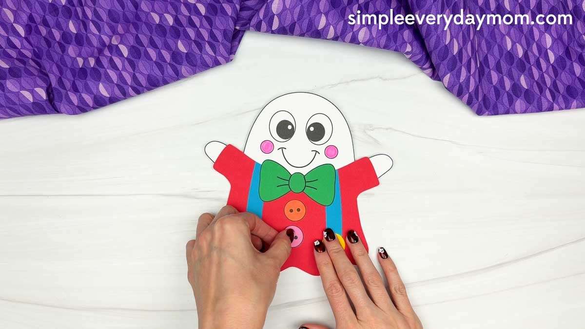 Hands attaching a pink button below the orange button on the red outfit of a clown ghost disguise craft.
