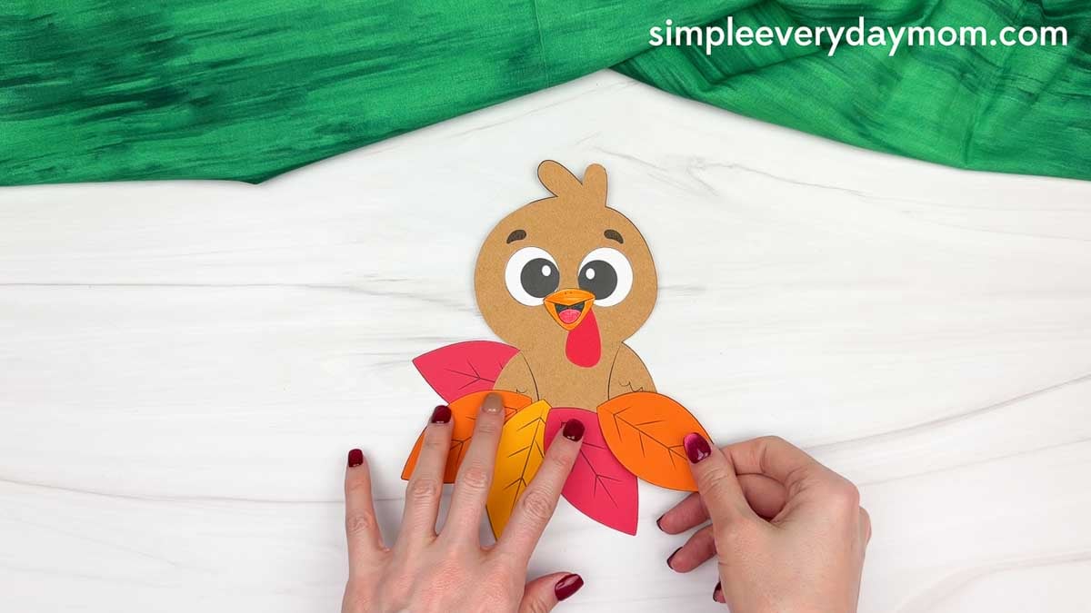 Hands arranging colorful autumn leaves along the bottom of a brown paper turkey for a fun fall turkey craft.