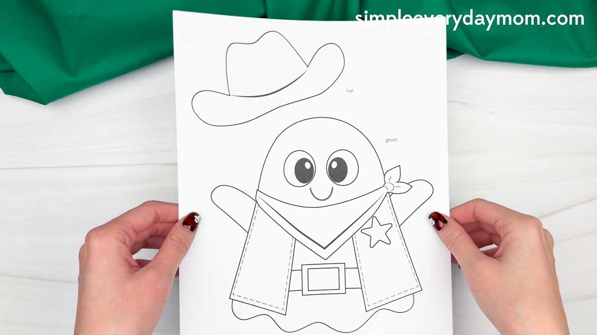 Hands holding a printable Cowboy Ghost Disguise Craft template featuring a ghost dressed in a cowboy hat, vest, bandana, and belt.