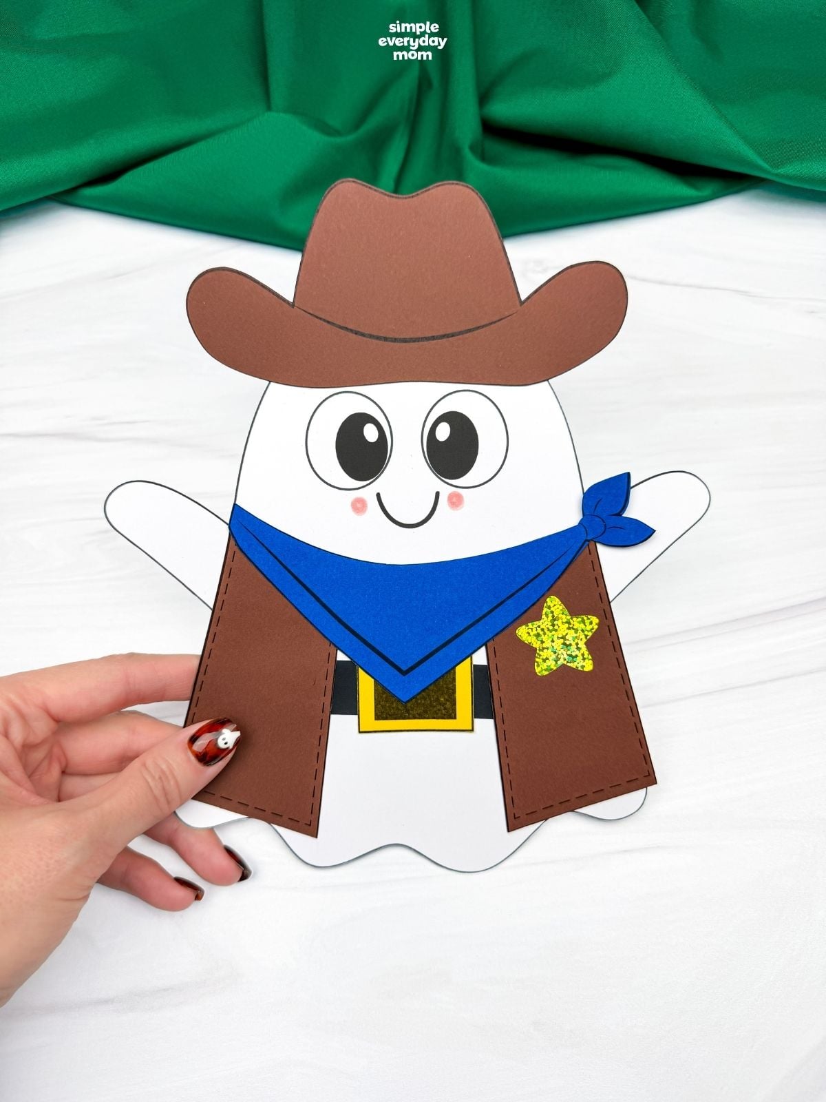 Hands holding a completed Cowboy Ghost Disguise Craft with a cowboy hat, bandana, vest, belt buckle, and star badge.