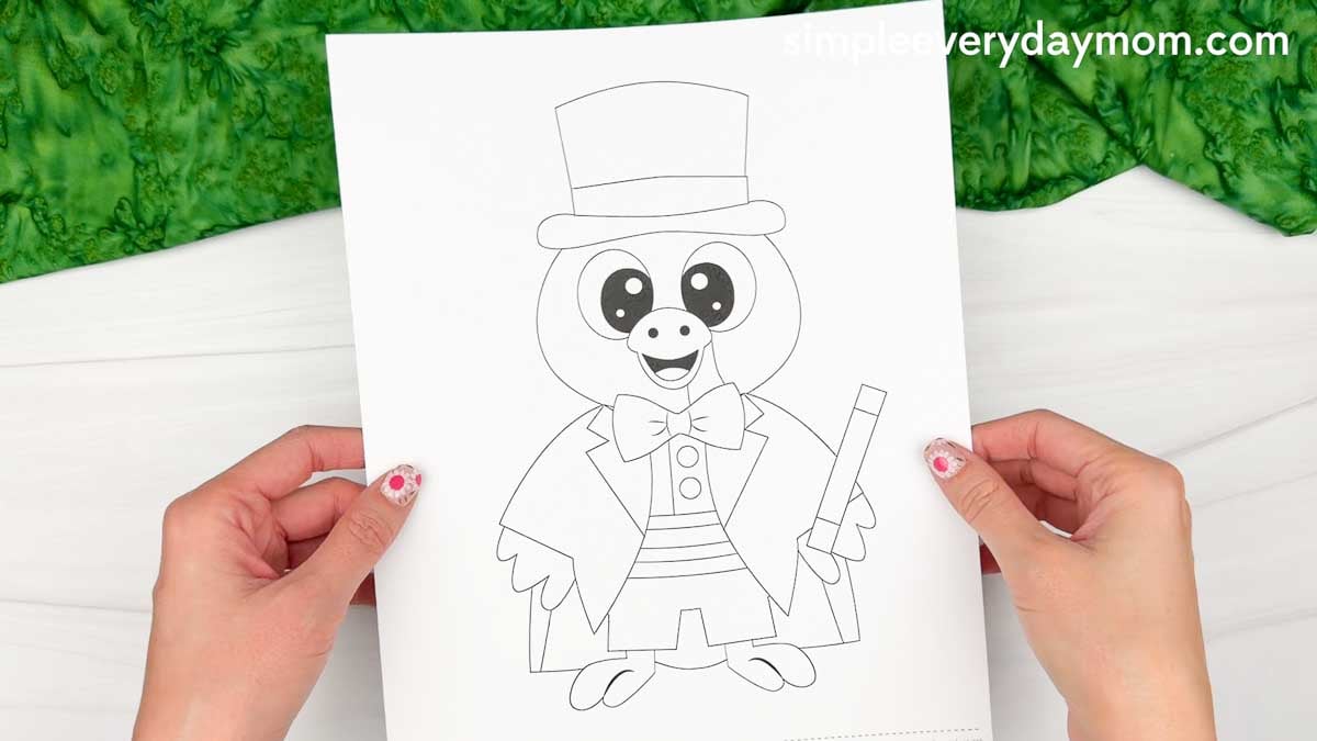 Hands holding a printable Magician Turkey Disguise Craft coloring page featuring a turkey wearing a top hat, bow tie, cape, and holding a magic wand.