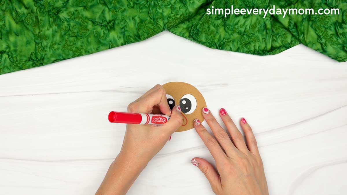 Hands drawing a red smile below the eyes on the brown turkey head using a marker for the Magician Turkey Disguise Craft.