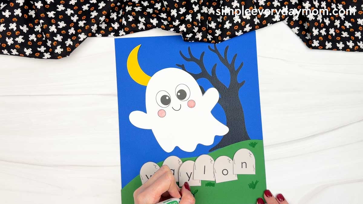 Hands adding grass details with a green marker below the lettered tombstones spelling a child's name for the Halloween Ghost Name Craft.
