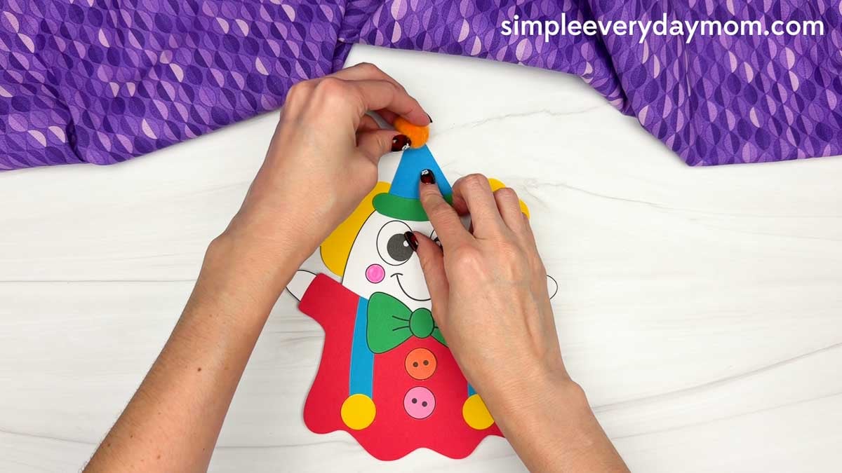 Hands attaching an orange pom-pom to the top of the blue party hat on a clown ghost disguise craft.