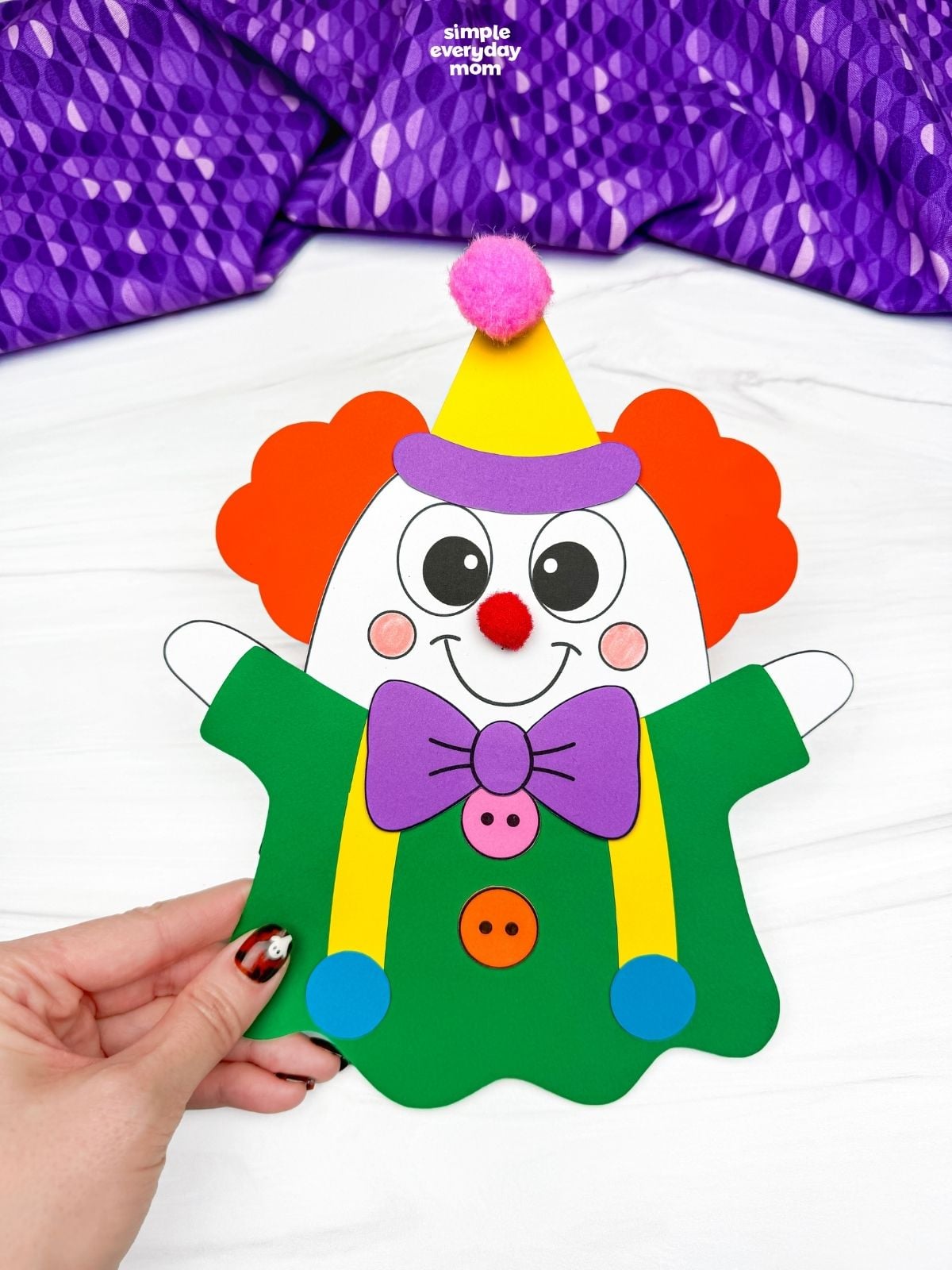 Hands holding a completed clown ghost disguise craft with orange hair, a yellow party hat, purple bow tie, and green outfit for a fun Halloween activity for kids.
