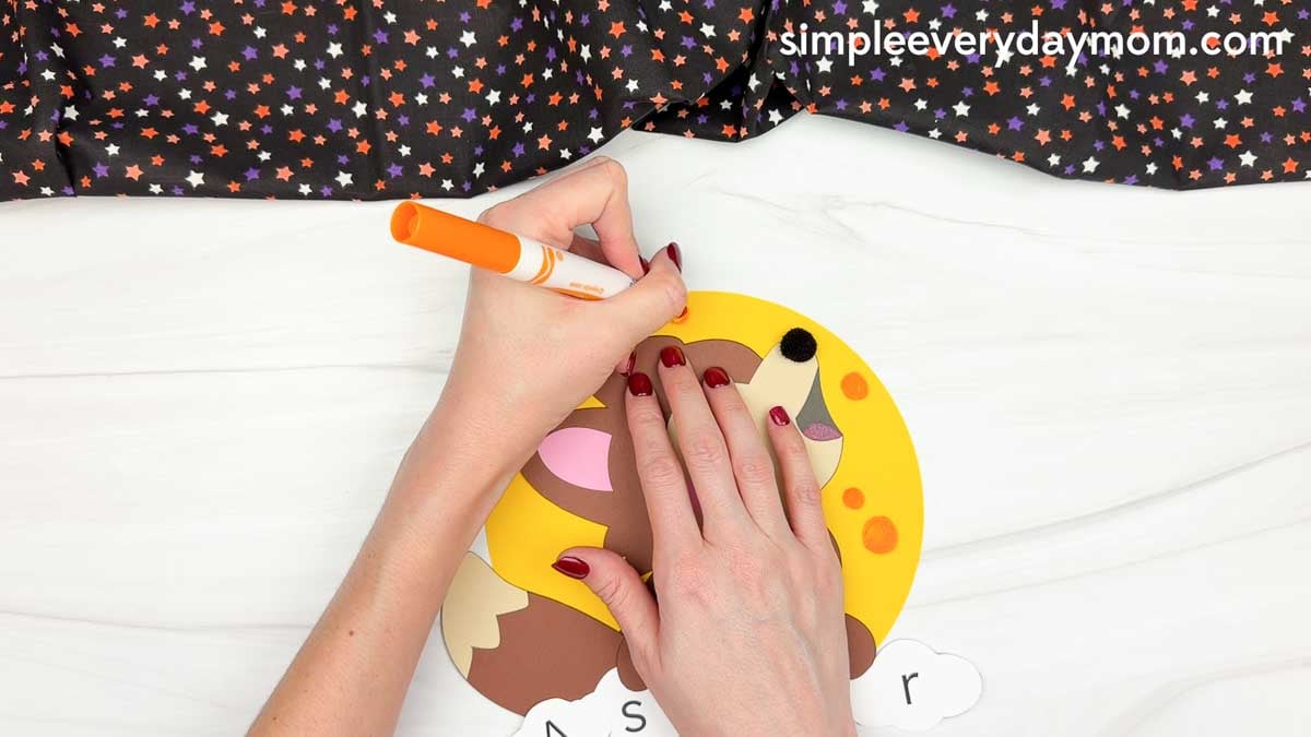 Hands using an orange marker to add circular spots to the full moon background of the werewolf name craft.