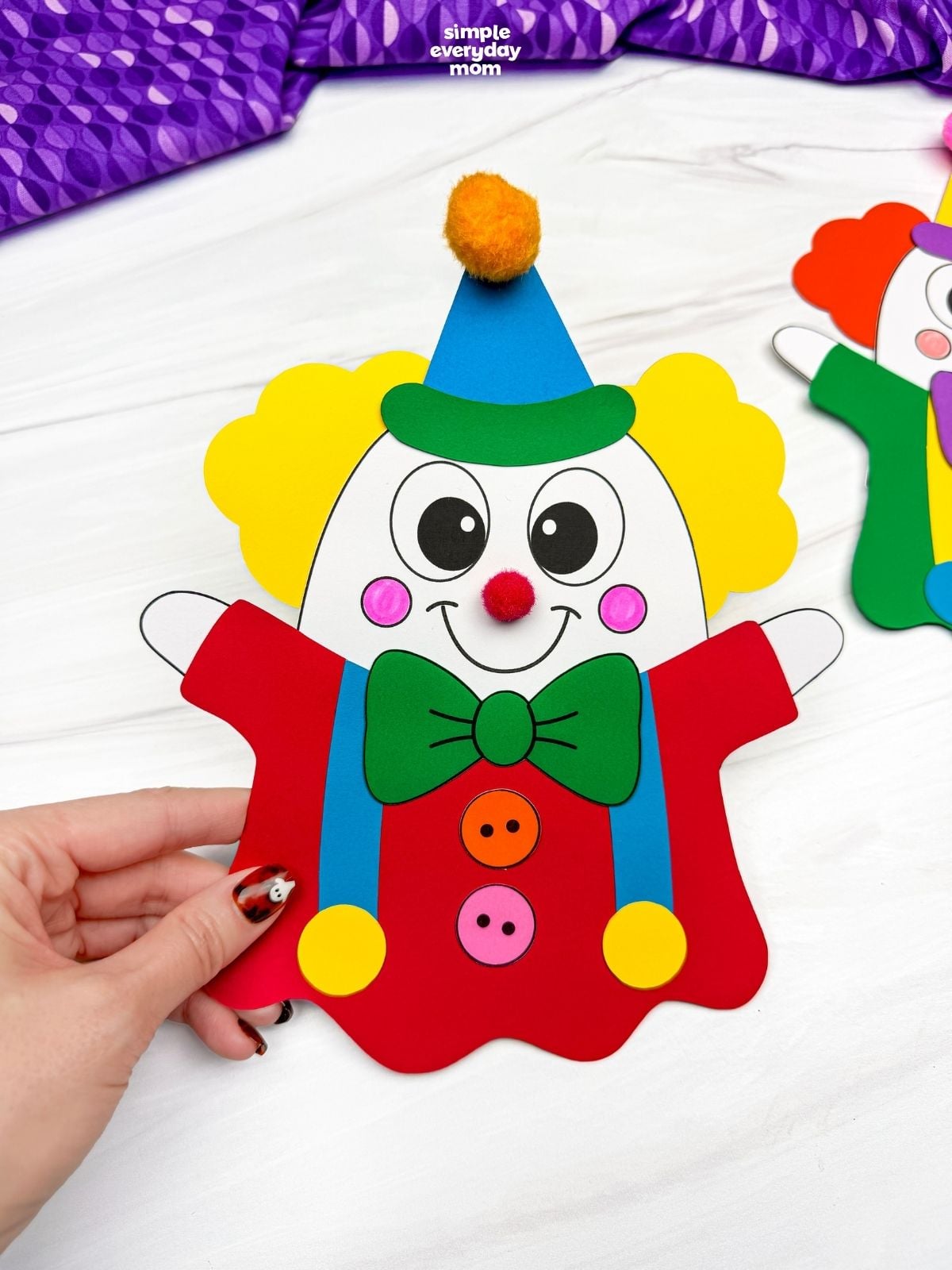 Hands holding a completed clown ghost disguise craft with yellow hair, a blue party hat, green bow tie, and red outfit for a fun Halloween activity for kids.