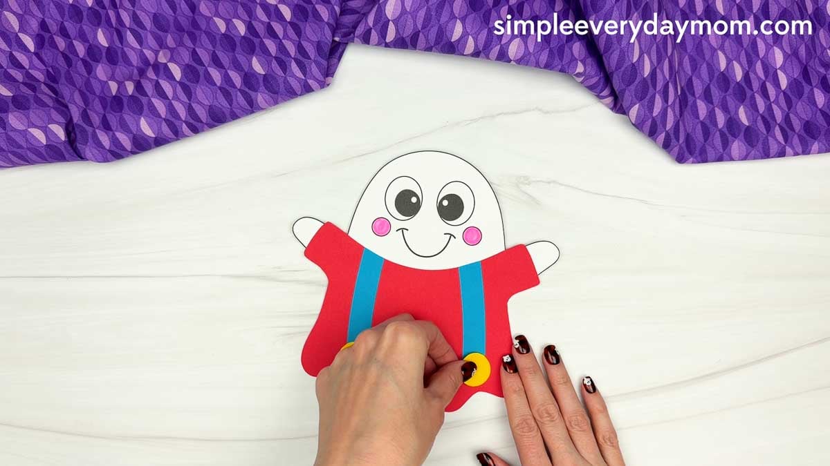 Hands attaching a yellow button to the blue suspenders on a red clown ghost disguise craft.