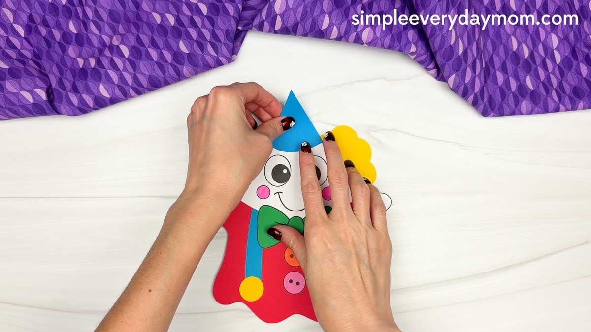 Hands attaching a blue party hat to the top of the clown ghost disguise craft between the yellow hair pieces.