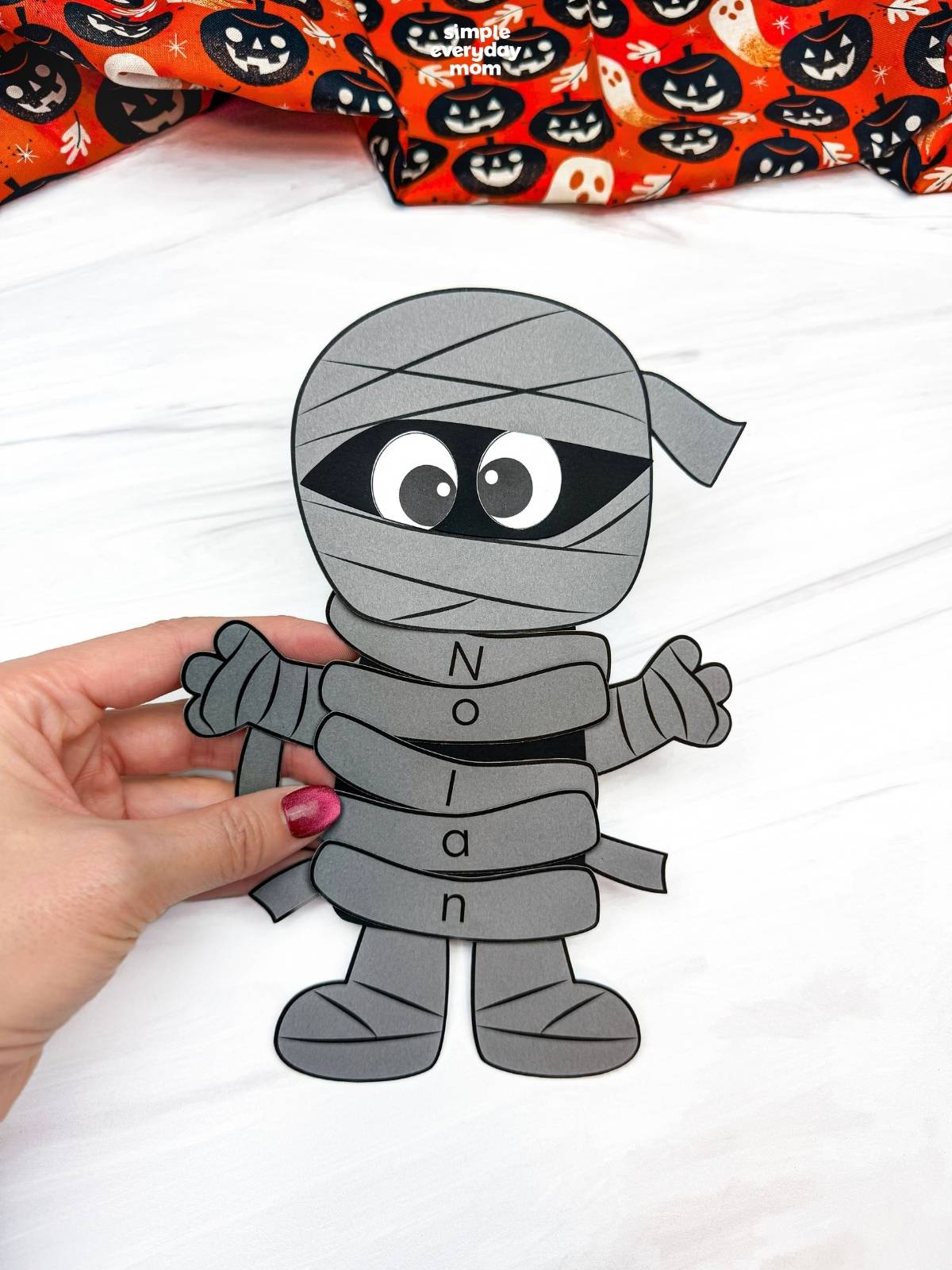 Hands holding a completed gray mummy name craft with lettered wraps spelling Nolan for a fun Halloween name recognition activity for kids.