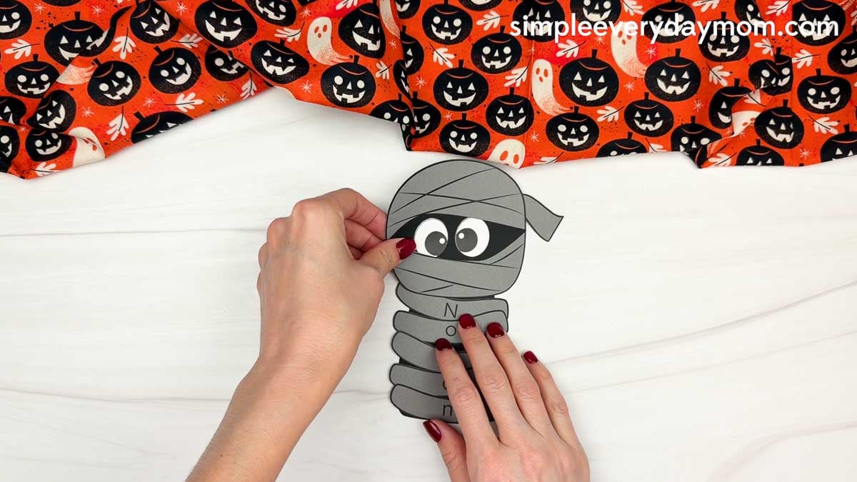 Hands gluing the mummy head above the lettered wraps on the body for a Halloween name craft for kids.