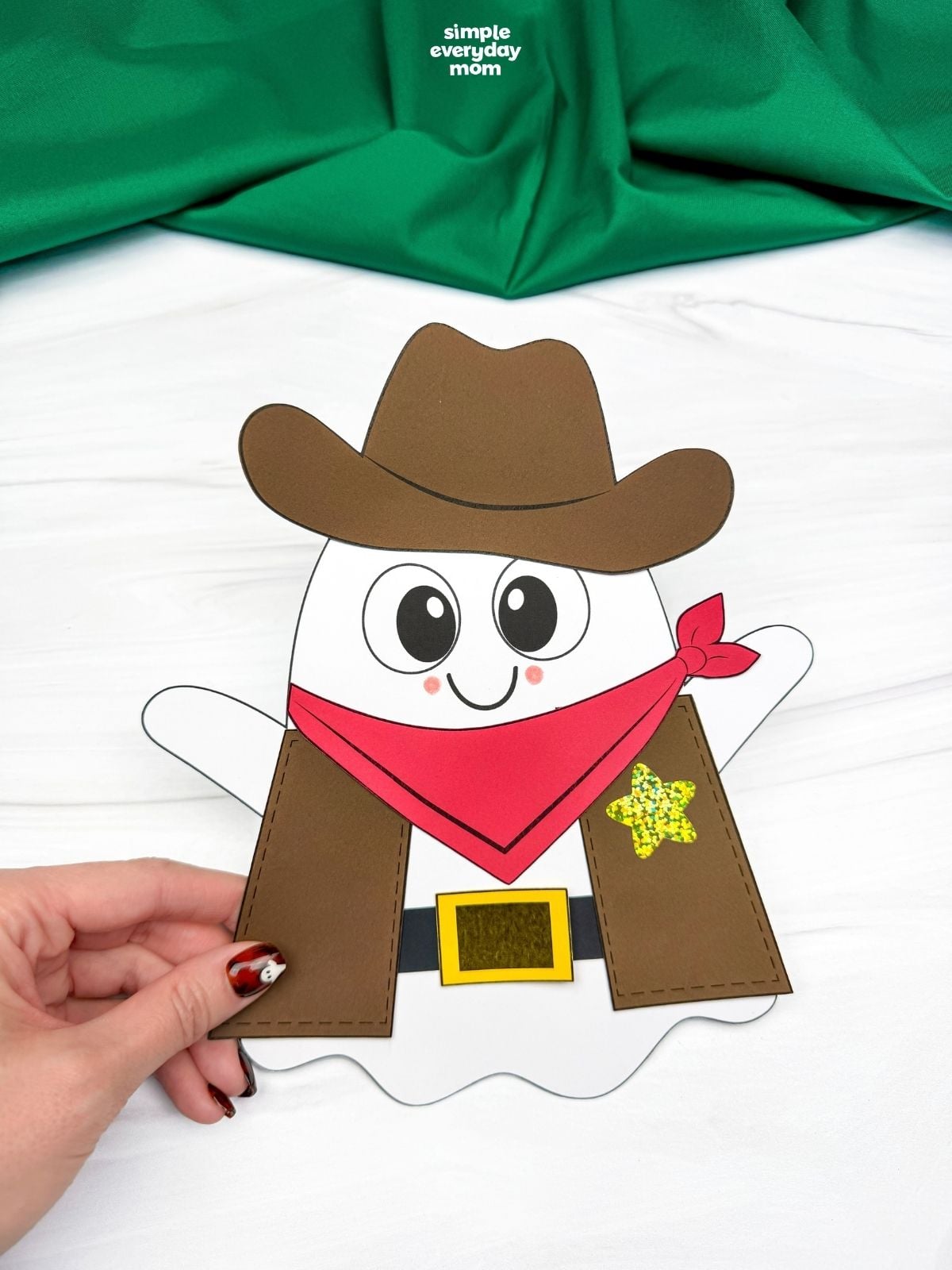 Hands holding a completed Cowboy Ghost Disguise Craft with a cowboy hat, bandana, vest, belt, buckle, and star badge.