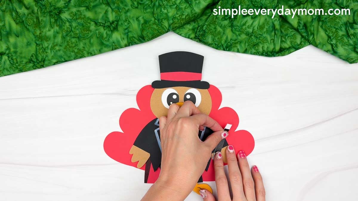Hands attaching a black magic wand with a white tip to the turkey's right wing for the Magician Turkey Disguise Craft.