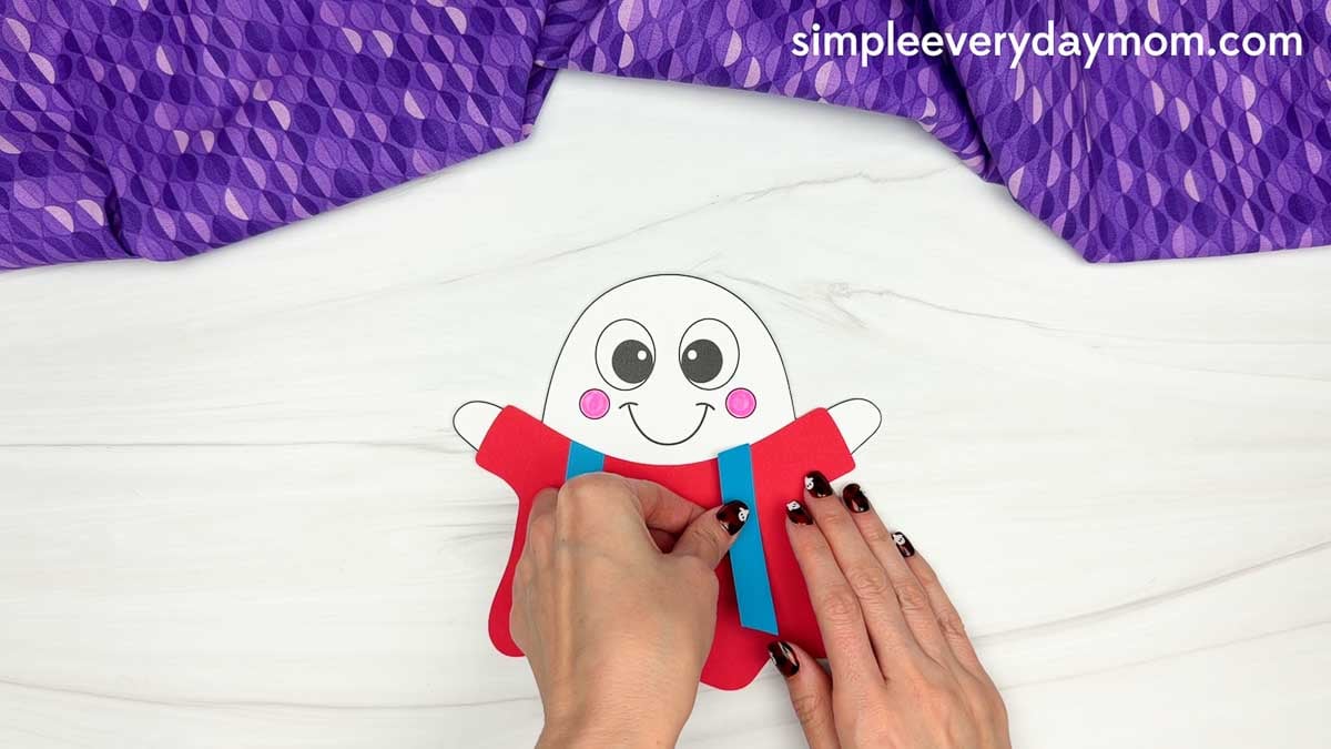Hands gluing blue suspenders onto the red body of a clown ghost disguise craft.