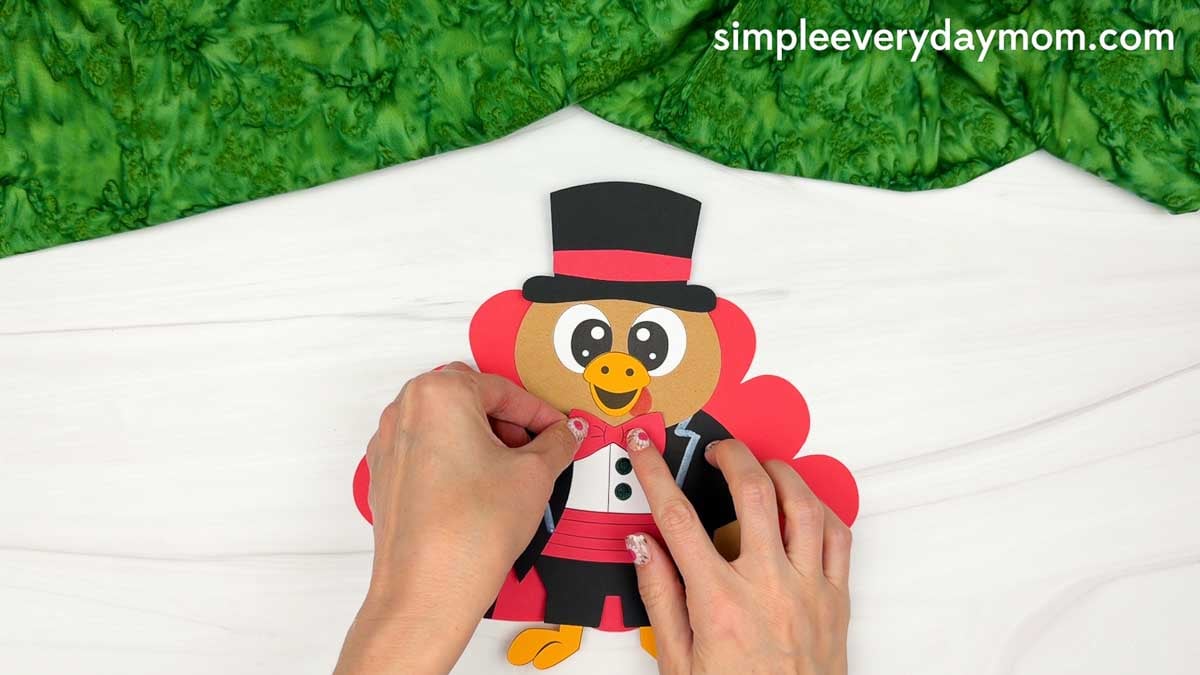 Hands attaching a red bow tie below the turkey's head, centered above the white shirt for the Magician Turkey Disguise Craft.