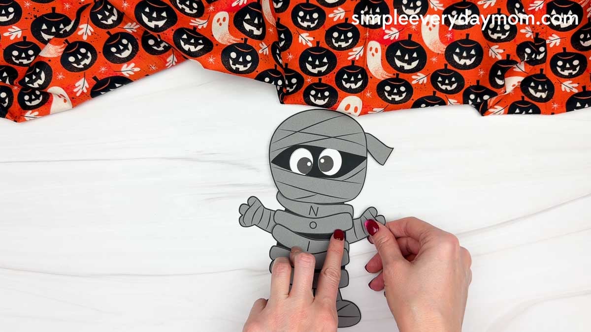 Hands attaching the remaining arm to the side of the mummy body for a Halloween name craft for kids.