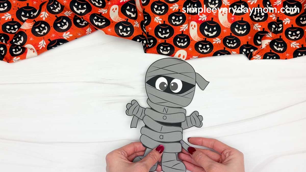 Hands adding an extra wrap to the lower body of the mummy name craft with lettered bandages for a Halloween activity for kids.
