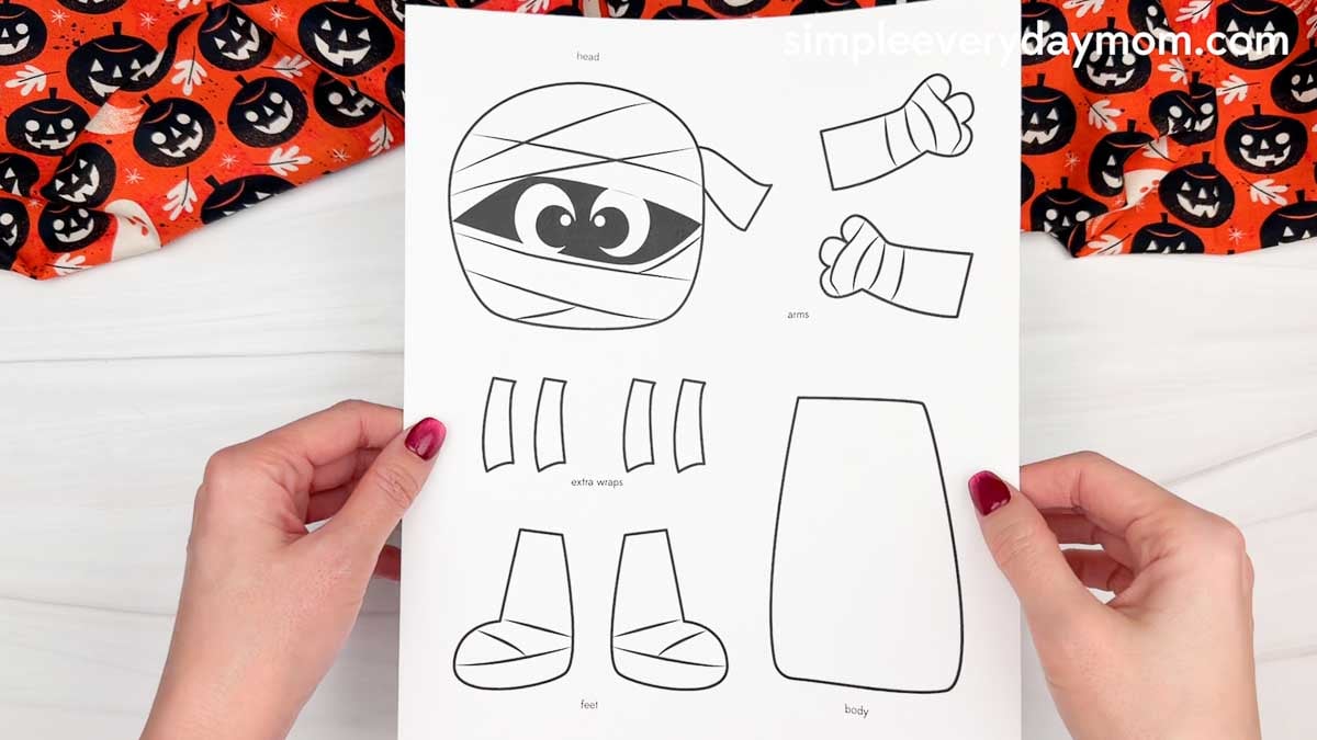 Hands holding a printable mummy name craft template featuring the head, arms, extra wraps, feet, and body pieces for a Halloween activity.