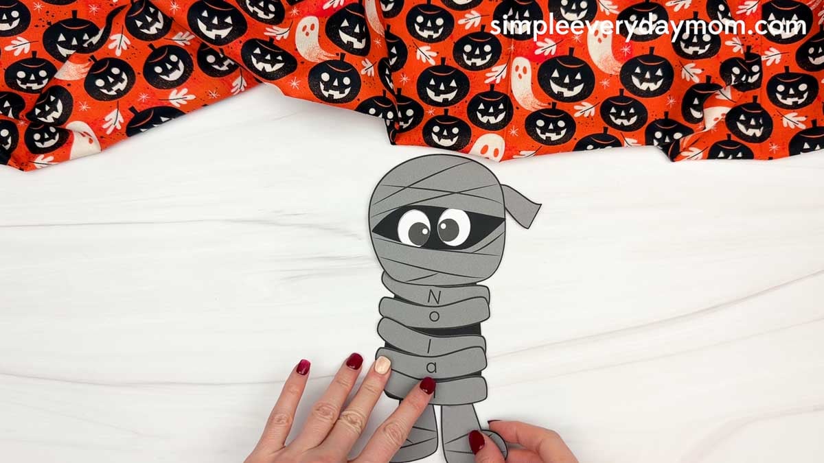 Hands attaching the feet to the bottom of the mummy body with lettered wraps for a Halloween name craft for kids.