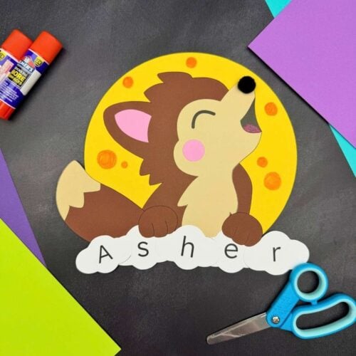 A completed craft featuring a howling werewolf against a yellow full moon with lettered pieces spelling a child's name, surrounded by Halloween paper craft supplies.