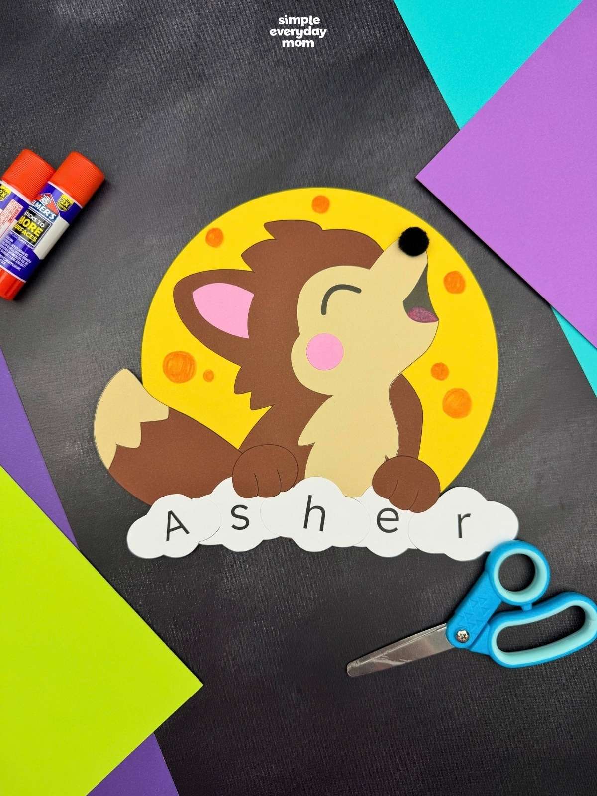 A completed craft featuring a howling werewolf against a yellow full moon with lettered pieces spelling a child's name, surrounded by Halloween paper craft supplies.