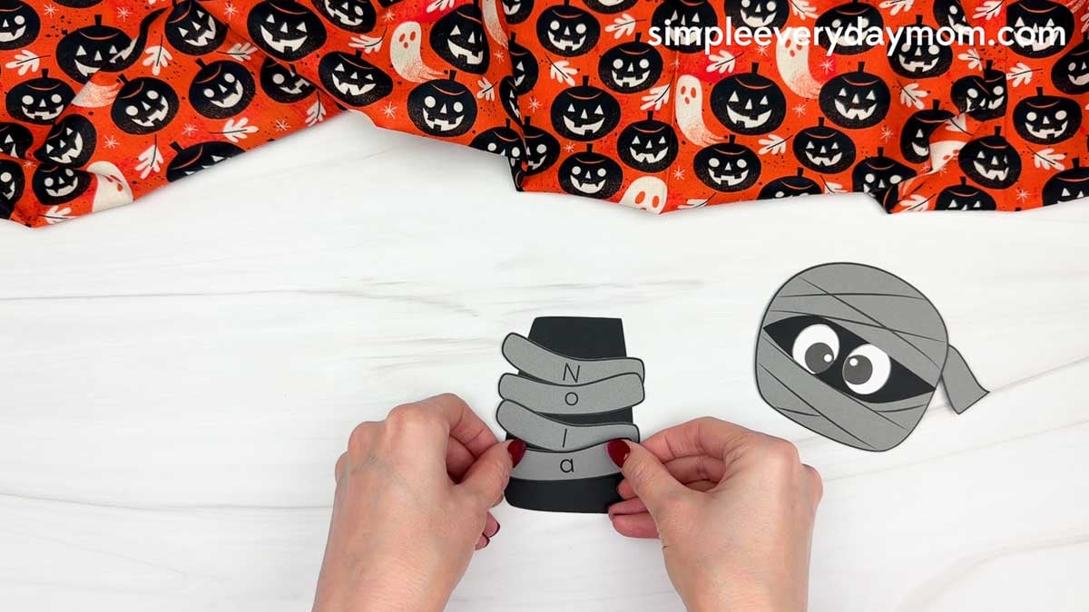Hands placing a lettered wrap onto the mummy body to spell a child's name for a Halloween name craft activity.