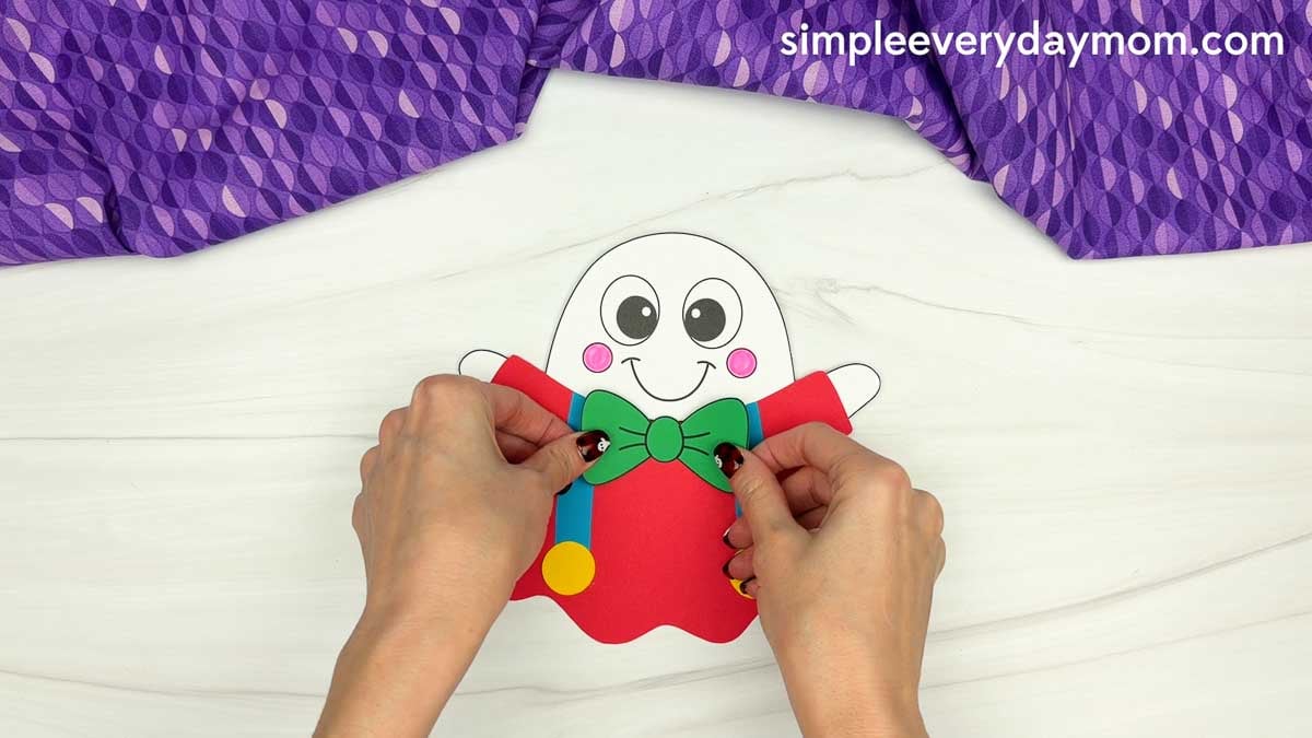 Hands placing a green bow tie below the face of a clown ghost disguise craft with red clothing and blue suspenders.
