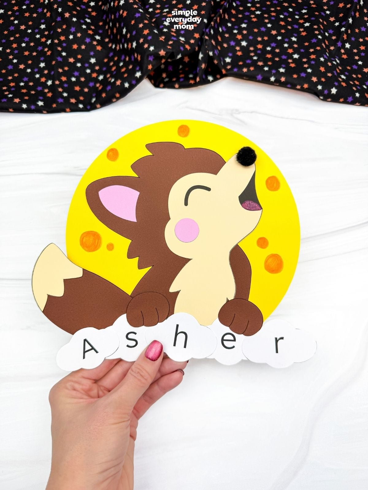 Hands holding a completed werewolf name craft featuring a howling wolf, yellow full moon, and lettered pieces spelling Asher for a fun Halloween activity.