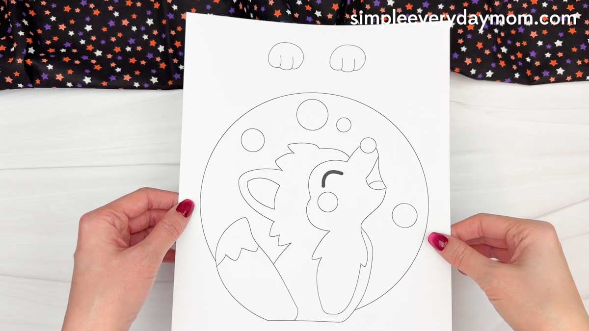 Hands holding a printable werewolf name craft template featuring a howling wolf, moon, and stars for a Halloween activity.