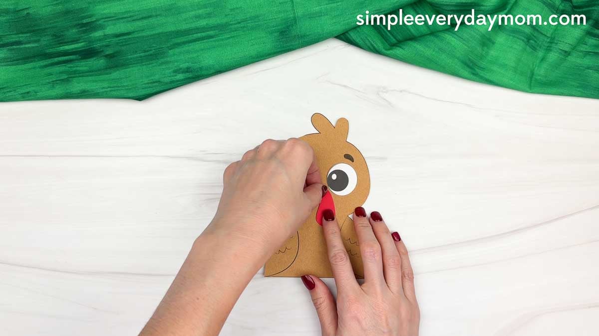 Hands attaching the turkey's red snood below the eyes on a brown paper fall turkey craft.