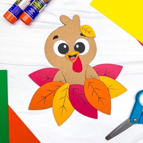 A completed craft featuring a cute brown paper turkey nestled in colorful fall leaves, surrounded by glue sticks, scissors, and construction paper.