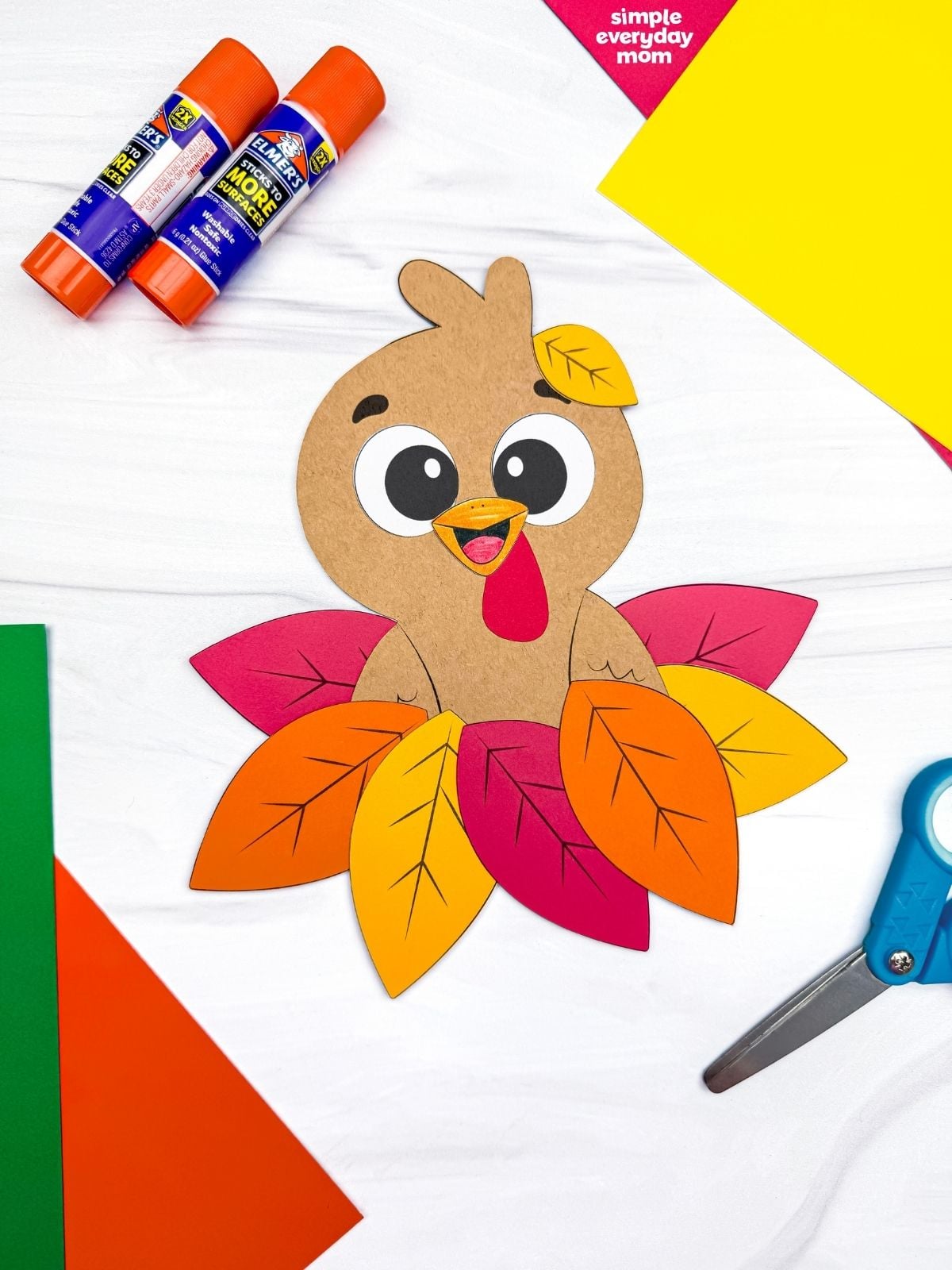 A completed craft featuring a cute brown paper turkey nestled in colorful fall leaves, surrounded by glue sticks, scissors, and construction paper.