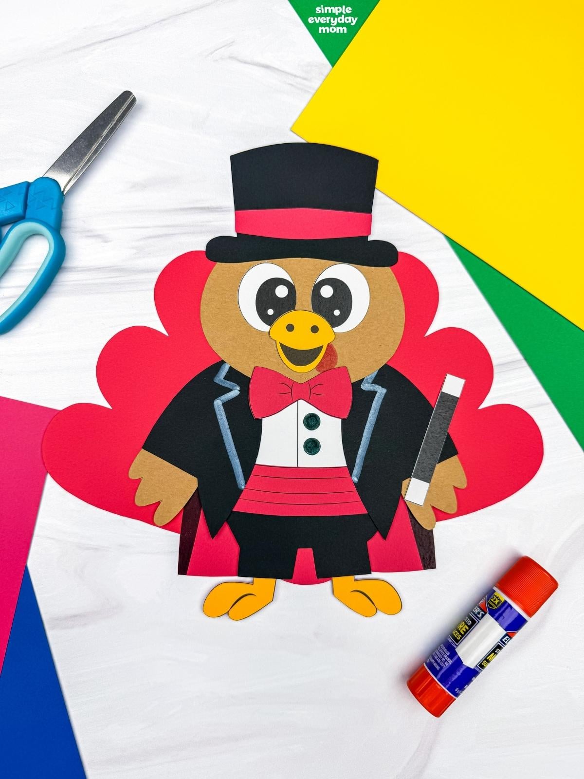 A completed craft of a magician turkey disguise featuring red tail feathers, a black top hat, red bow tie, and magic wand, surrounded by colorful craft supplies.